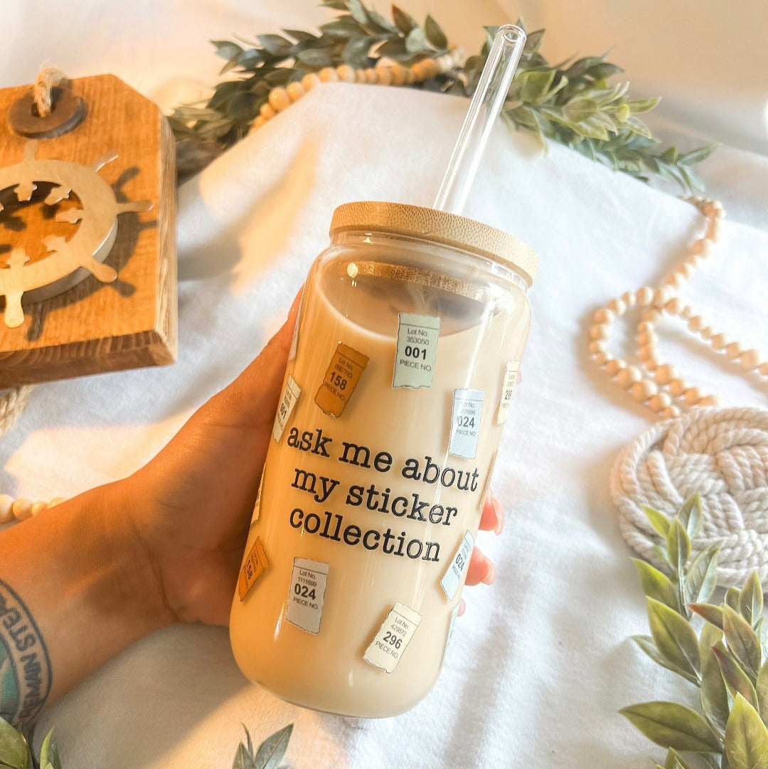 A hand holds a clear glass can cup with a bamboo lid and clear straw, filled with a light tan beverage. The jar is decorated with small PCS stickers and the text 'ask me about my sticker collection'.